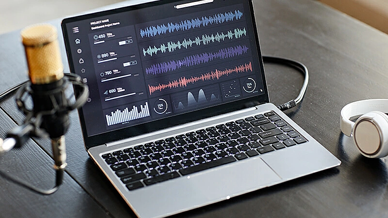 Imagem de um microfone focado e ao fundo uma tela de notebook com ondas de áudio.