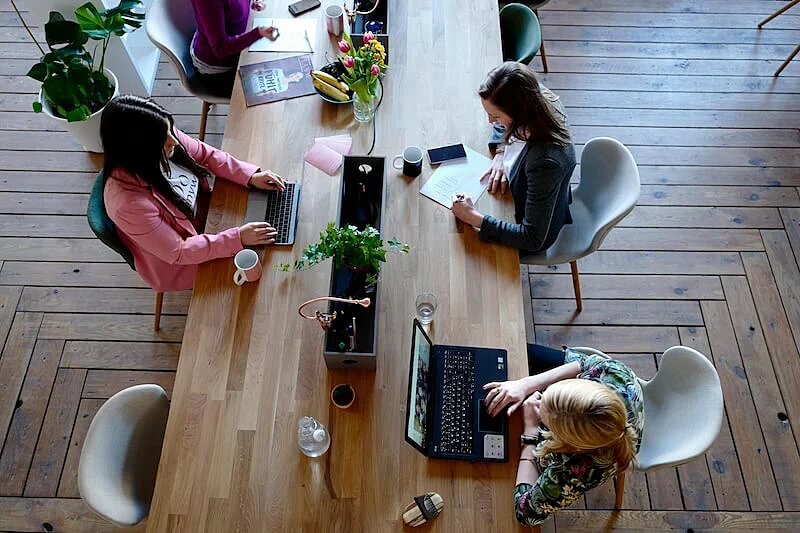 The image shows four women working in an office. They are sat on a table, two of them are using a laptop, an the other two are reading a paper. 