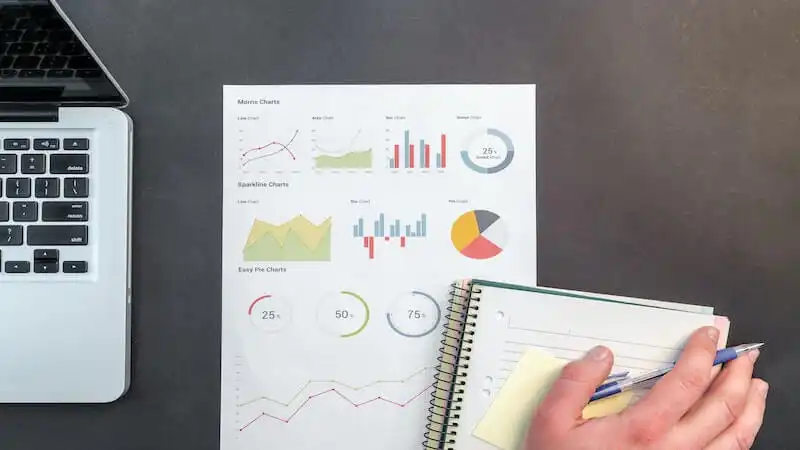 The image shows a paper where there are many graphics, on the left side there is a laptop, and on the inferior right side of the image there is a hand holding a pen, above a notebook